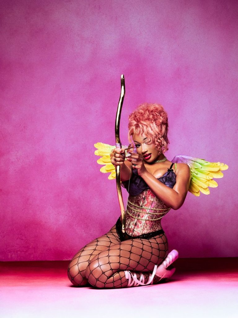 Megan Thee Stallion sit on her knees and is aiming a bow at the camera