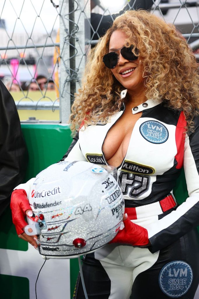 Frontal view of Beyonce standing more to the side and smiling while wearing a racing outfit and holding a helmet