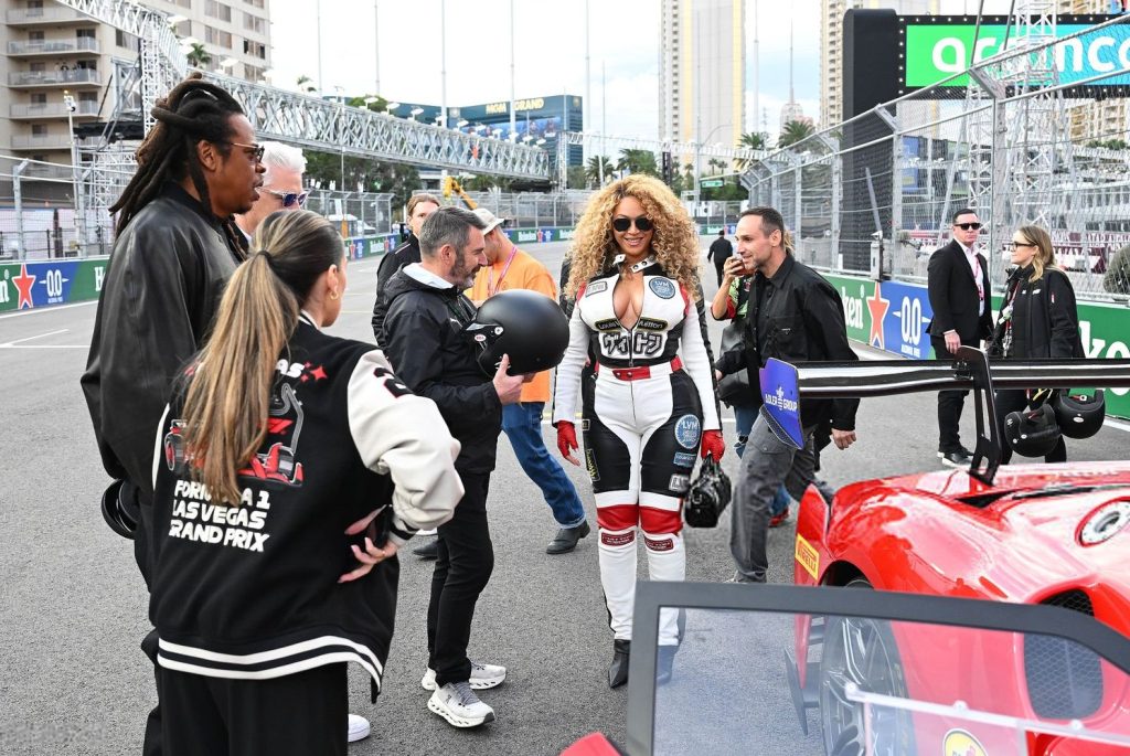 Horizontal full body frontal view of Beyonce in a racing outfit and standing in the middle of a group of people next to a racing car