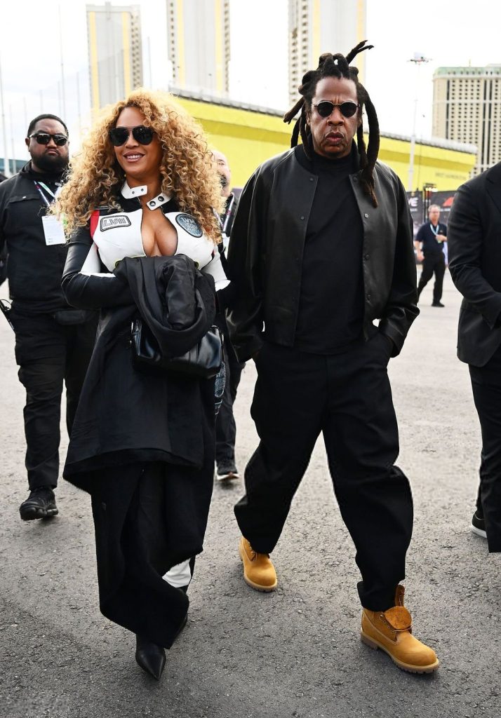 Full body frontal of Beyonce wearing a racing outfit and looking to the side while walking next to Jay-Z