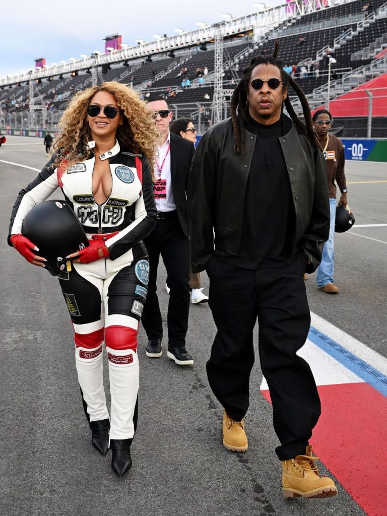 Full body frontal of Beyonce in a racing outfit and holding a helmet while walking next to Jay-Z