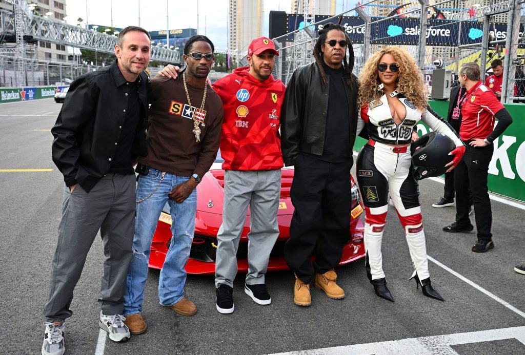 Horizontal full body view of Beyonce, Jay-Z, Lewis Hamilton and others posing in front of a racing car