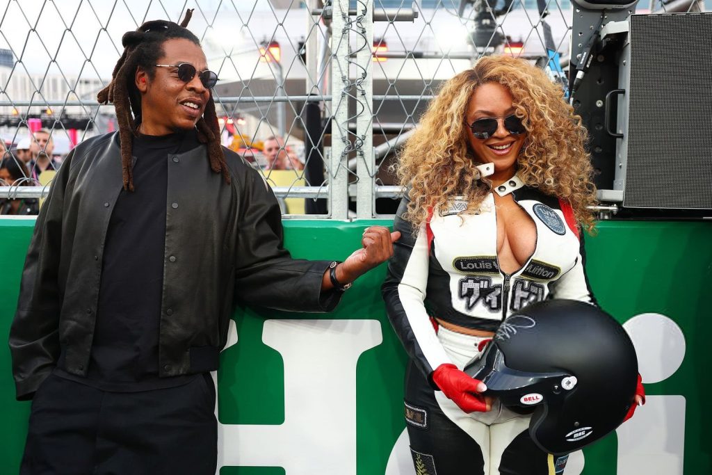 Close up of Beyonce holding a helmet and wearing a racing outfit while standing next to a smiling Jay-Z