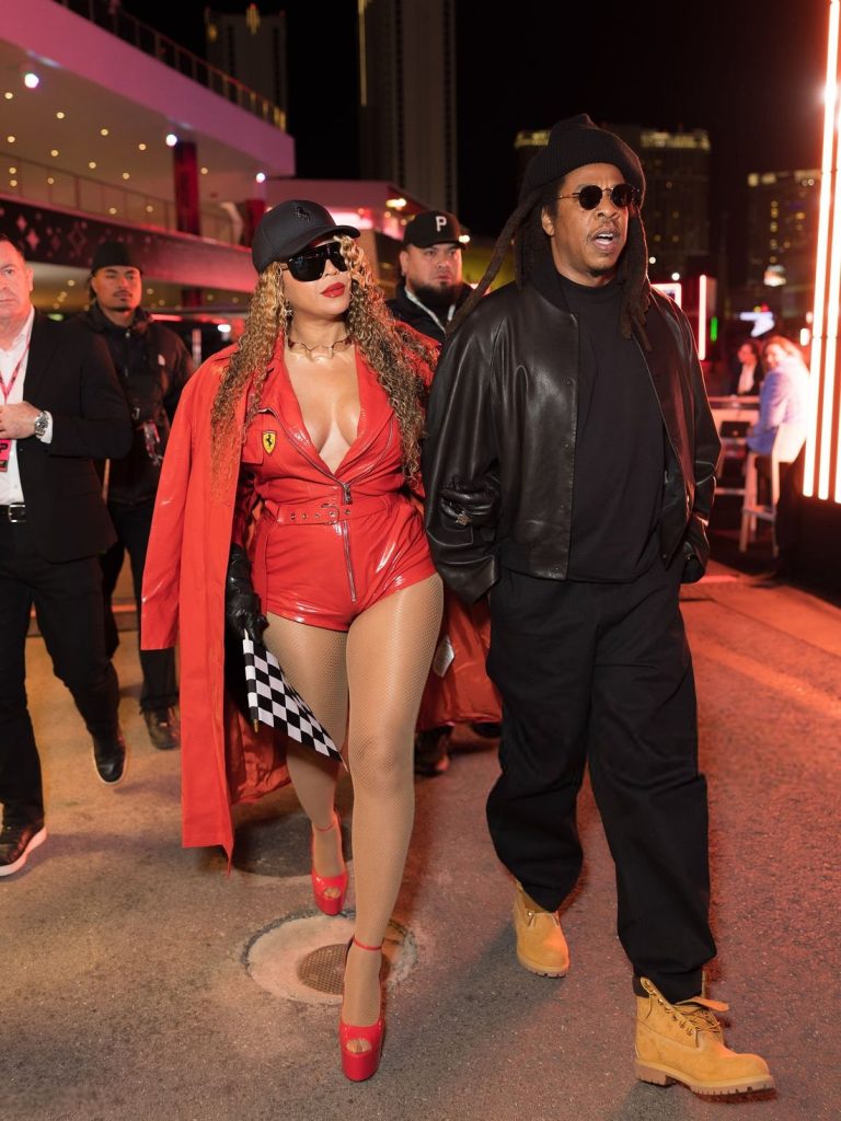 Full body frontal view of Beyonce walking next to Jay-Z wearing a red leather Ferrari outfit