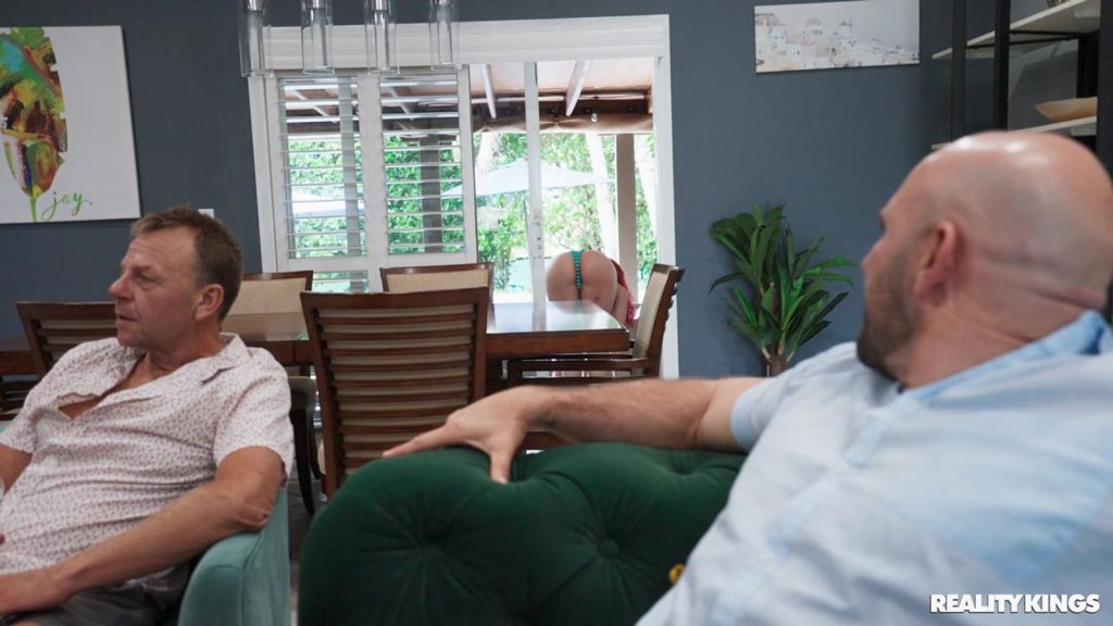 Two men sitting on couches and one of them is looking back to look at a girl bending over in a bikini