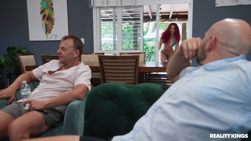 Two men sitting on couches and one of them is looking back to look at a girl standing up in a bikini