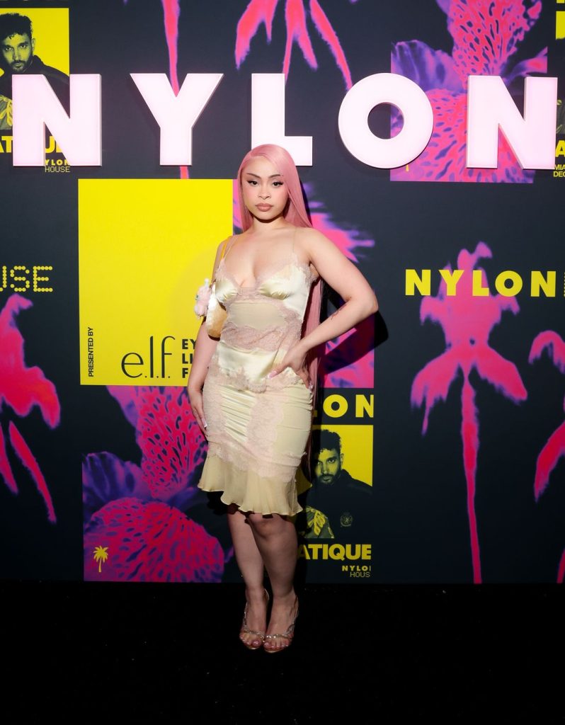 Far away frontal view of Ice Spice posing in a sexy dress on a red carpet with the word NYLON written in the background
