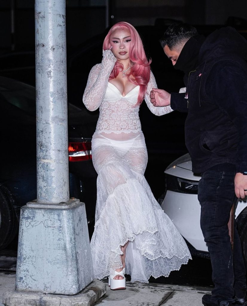 Full body frontal of Ice Spice looking into the camera as she is getting help up a curb