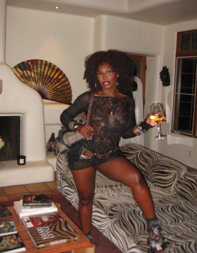 A woman in a black lace dress poses indoors with one leg on a zebra-striped couch, holding a glass of wine. The room has eclectic decor, including a fan wall ornament, books, and patterned textiles.