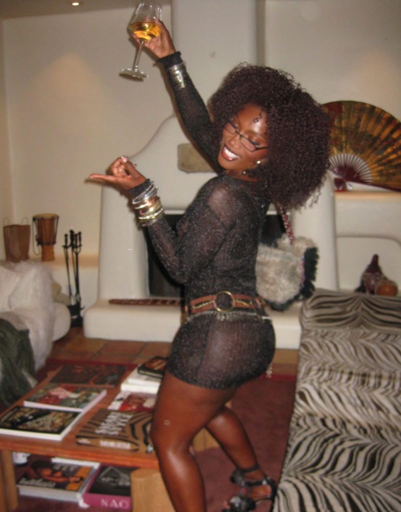 A woman wearing a sheer black dress, high heels, and jewelry poses playfully with a drink in hand, smiling in a stylish living room with animal print decor and coffee table books.