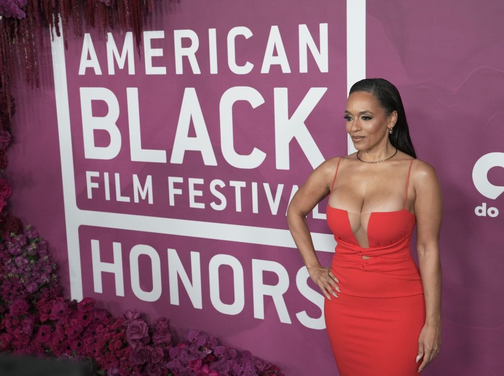 A woman in a red dress poses in front of a magenta backdrop with white text that reads American Black Film Festival Honors, surrounded by purple and pink flowers.