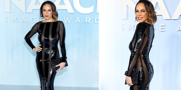 A woman poses on the red carpet in a long-sleeved, black sequin dress with sheer side panels at an awards event, smiling with red lipstick and styled hair, holding a black clutch.
