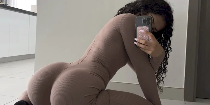 Fit girl wearing a fitted, long-sleeve brown jumpsuit kneels on a white tiled floor, holding a phone up to cover her face.