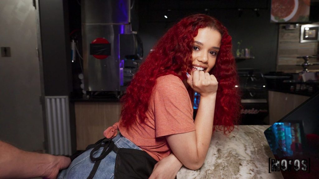 A young woman with long, curly red hair smiles shyly, resting her chin on her hand. She wears a pink t-shirt and denim apron, leaning on a counter in a dimly lit restaurant or café setting.