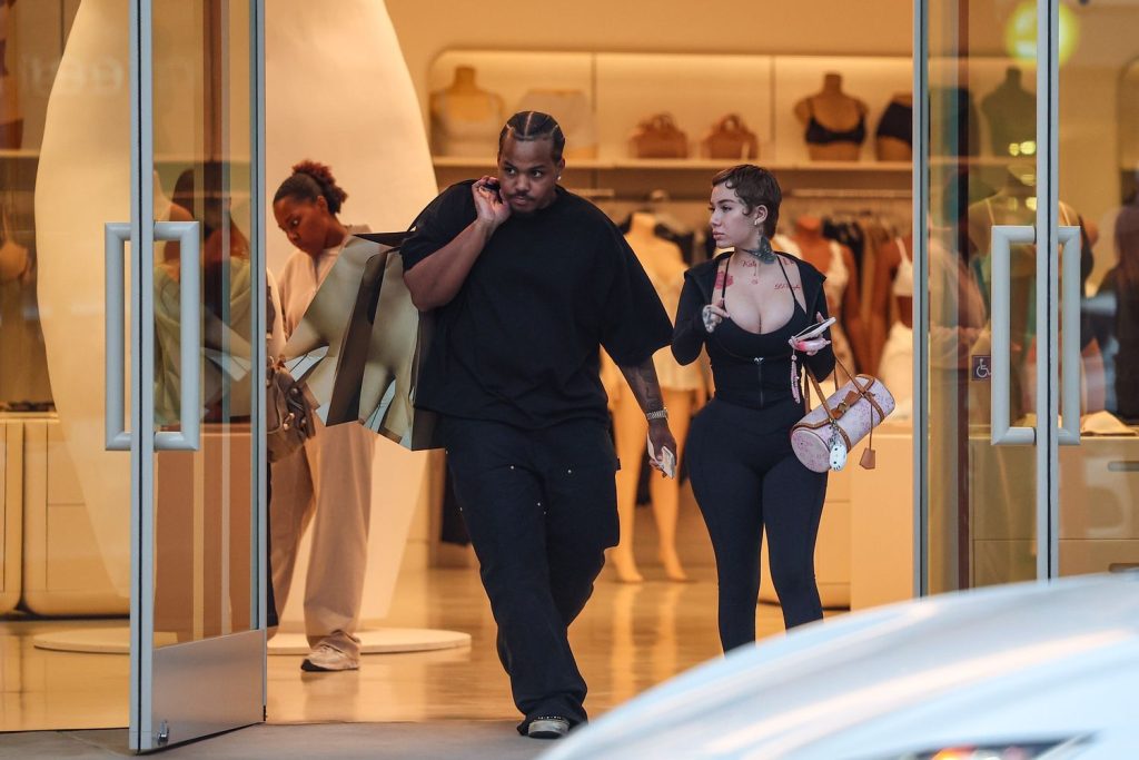 A man and woman exit a clothing store carrying shopping bags. The man wears a black shirt and pants, holding several bags, while the woman wears a black outfit and carries a small purse. Store displays and shoppers are visible inside.