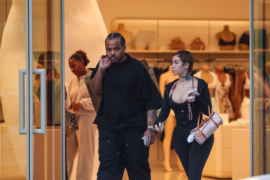A man and a woman walk out of a clothing store. The man is dressed in black and talks on the phone, while the woman, also in black, carries a pink bag and holds his arm. Mannequins and clothing racks are visible inside the store.