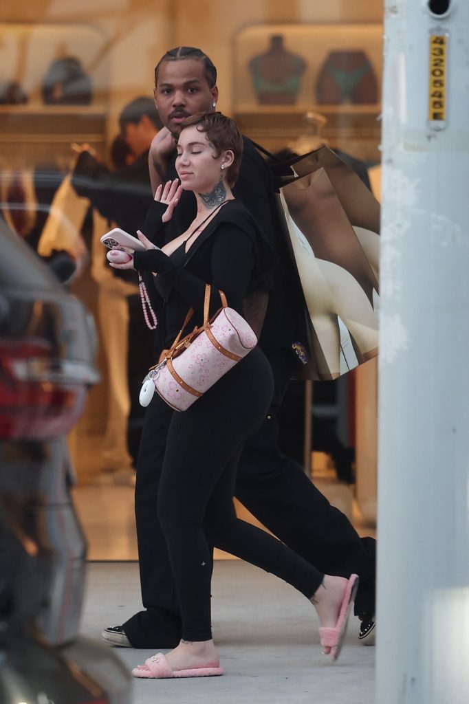 A woman in black clothes and pink slippers walks beside a man in a black outfit, carrying shopping bags. She has short hair, visible tattoos on her neck, and a pink designer bag. Store displays are visible in the background.
