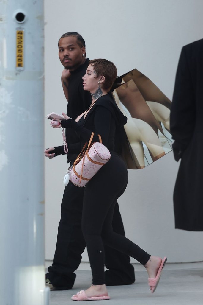 A woman in black leggings, pink slides, and a pink bag walks with a man dressed in black. She carries a shopping bag with a nude-figure design. They are outdoors, walking past a white wall and a metal post.