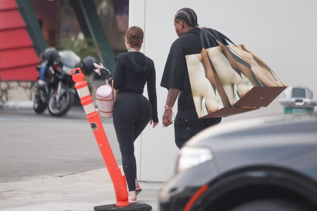 A woman in black leggings walks beside a man carrying large shopping bags with nude images printed on them. They are outdoors near a sidewalk with a traffic cone and a parked motorcycle in the background.