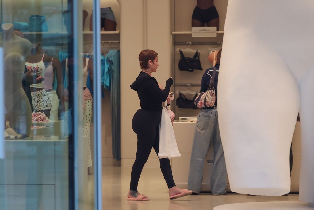 A woman in black leggings and a hoodie shops inside a clothing store, holding a white tote bag. Another person stands at the counter. Mannequins and folded clothes are visible in the background.