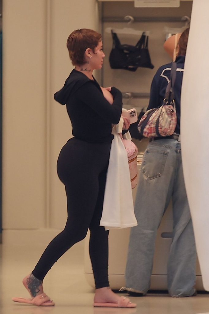 A woman with short hair and tattoos, dressed in a black hoodie and black leggings, stands indoors holding a white towel. She is wearing pink sandals and appears to be talking to another person at a counter.