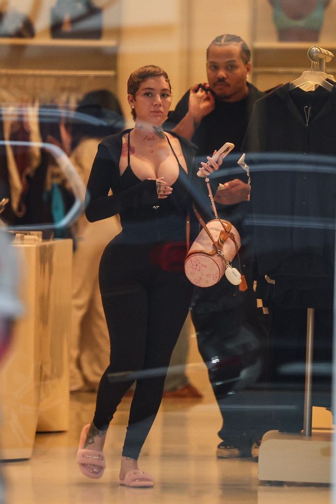 A woman in a black outfit and pink slippers holds a designer bag and phone while walking through a clothing store, with a man in dark clothing walking behind her. Clothing racks are visible in the background.