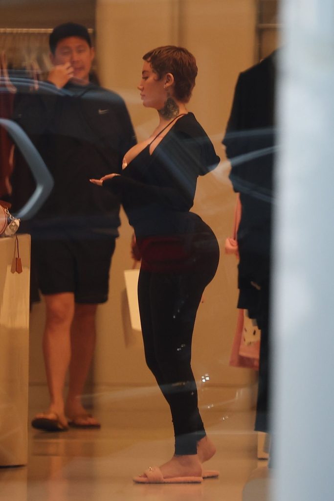 A woman with short hair, tattoos, and dressed in black leggings and a top stands in a retail store, facing a counter. Another person in casual clothing stands in the background.