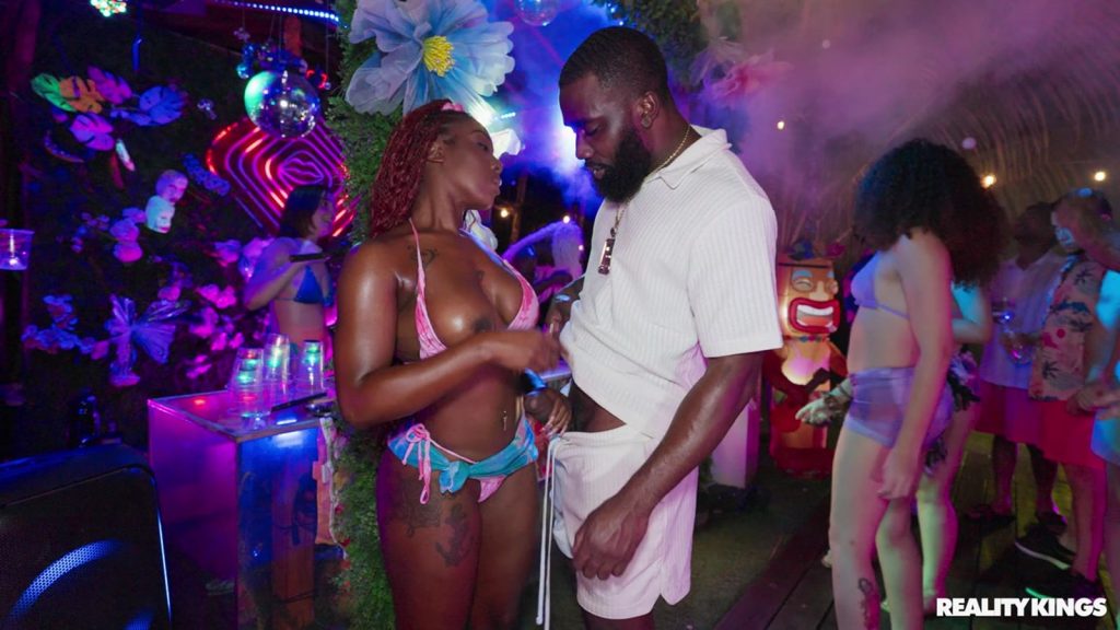 Two people stand close, getting intimate by helping each other undress at a lively, colorful party with neon lights, flowers, and dancing guests in swimwear in the background.