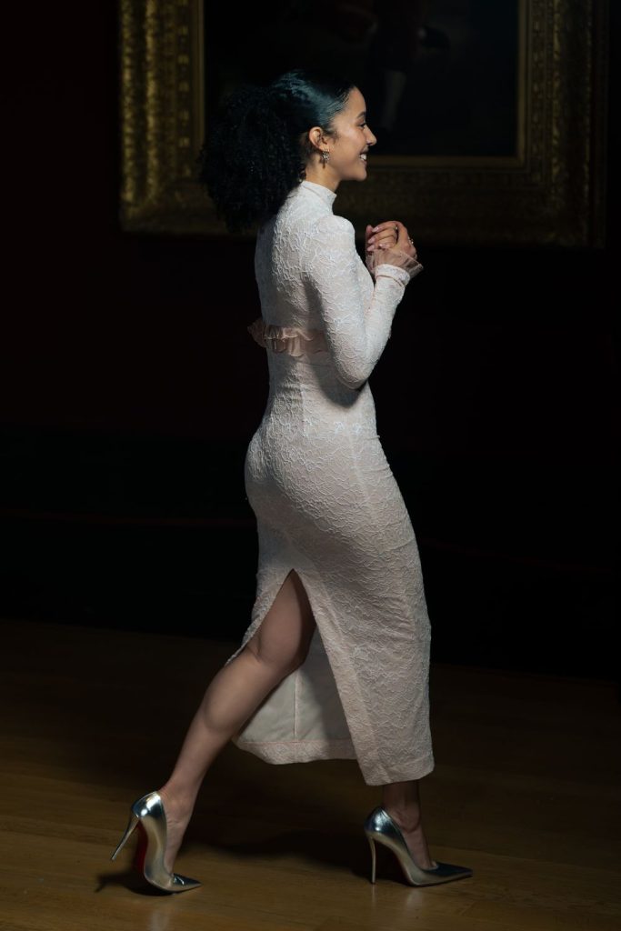 A woman in a long-sleeved, light pink lace dress and silver high heels walks indoors, smiling with her hands clasped. She is seen in profile against a dark background with a framed painting.