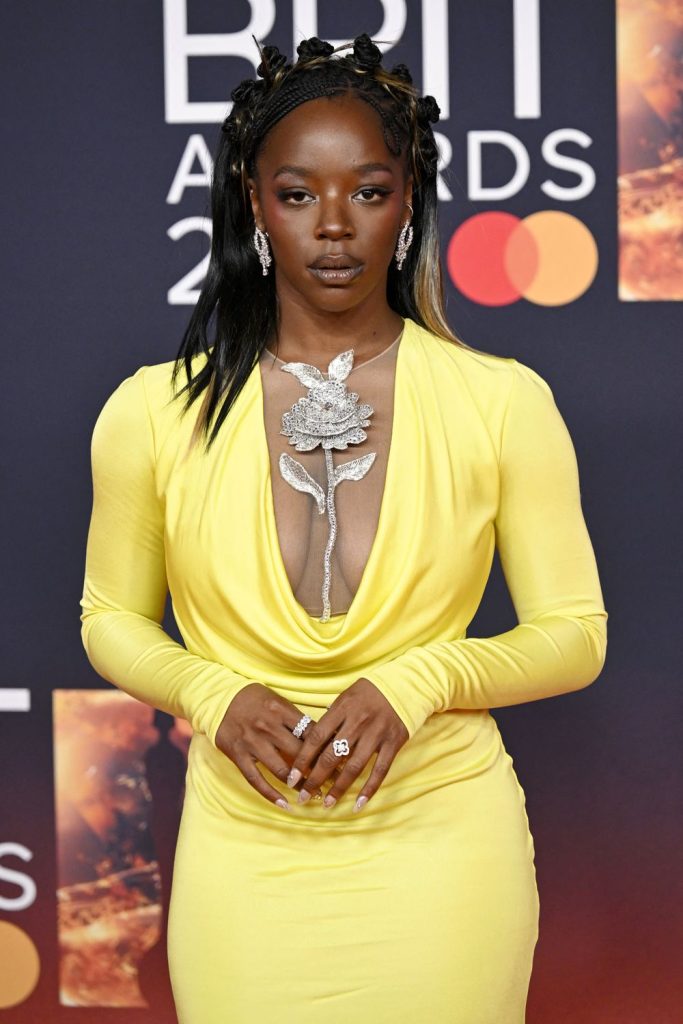 A black woman wearing a bright yellow, deep V-neck gown with a large silver flower necklace poses on the red carpet at the BRIT Awards. She has long braided hair, statement earrings, and dramatic makeup.