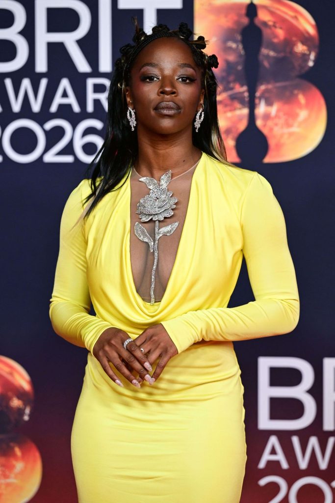 A black woman poses on the red carpet at the BRIT Awards 2024, wearing a bright yellow long-sleeved dress with a deep neckline and a large silver floral necklace, standing in front of a BRIT Awards backdrop.