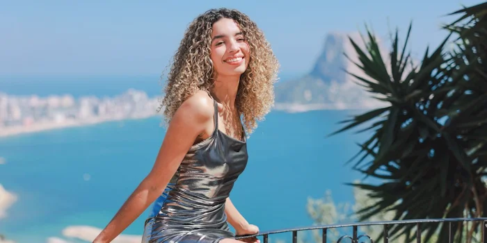 A girl with curly blonde hair smiles as she sit on the railing of a lookout point. She is wearing a shiny silver dress. Ocean and the outline of a bay can be seen below her in the background.