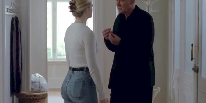 A young woman with blonde hair in a bun and a white top stands facing an older man in black clothing, who is gesturing with his hands as they talk in a bright, white room with large windows and a coat rack.