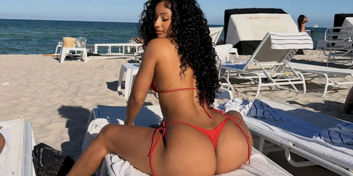 Rear view of a black woman with long curly hair wearing a red bikini sits on a beach lounge chair. The beach is sunny with clear blue sky, and empty lounge chairs are visible in the background.