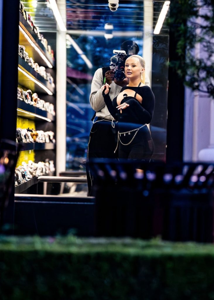 From the outside looking through the window of a shoe store. A blonde girl wearing black turns around to look out the window. A man with a big camera stands behind her.