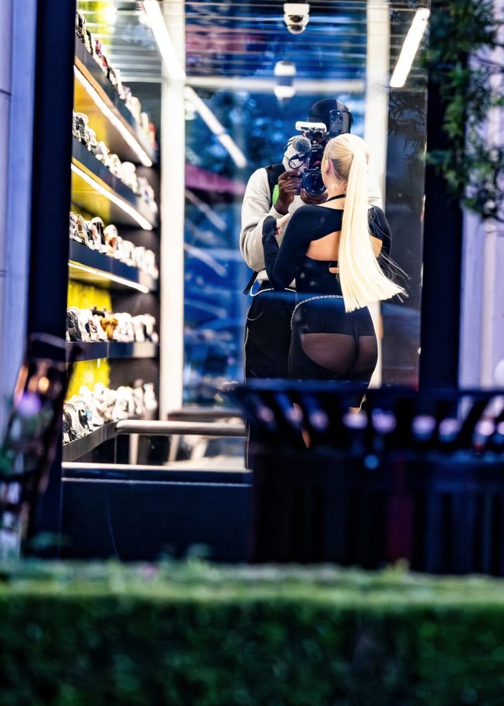 From the outside looking through the window of a shoe store. A blonde girl wearing semi-sheer black shorts face a man filming her with a big camera stands behind her.