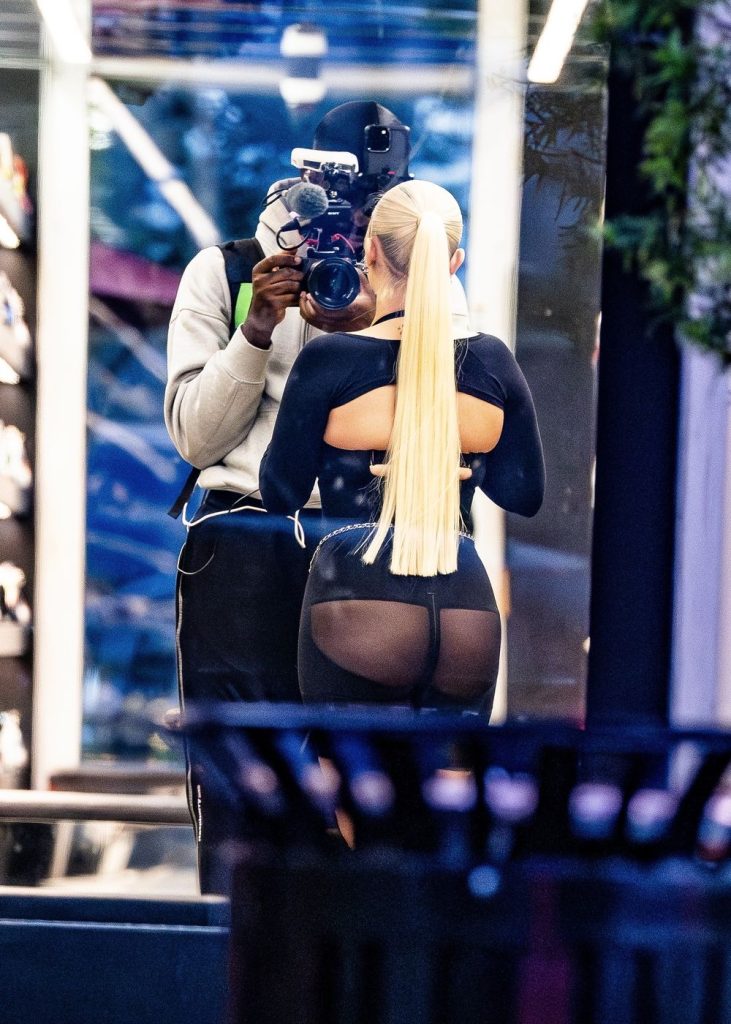 Close up of girl wearing semi-sheer black shorts facing away and standing in front of a man filming her with a big camera.