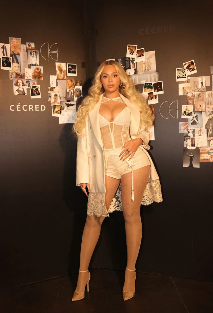 Beyonce standing in front of a brown wall with pictures on it wearing a corset-style top and a jacket.