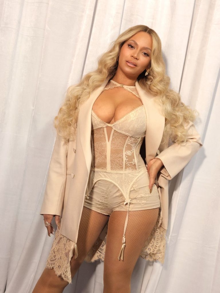 Beyonce standing with a hand on her hip and looking into the camera, wearing a corset style top in front of white curtains.