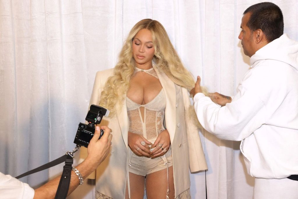 Beyonce dressed in white getting help with her hair and a camera is visible.