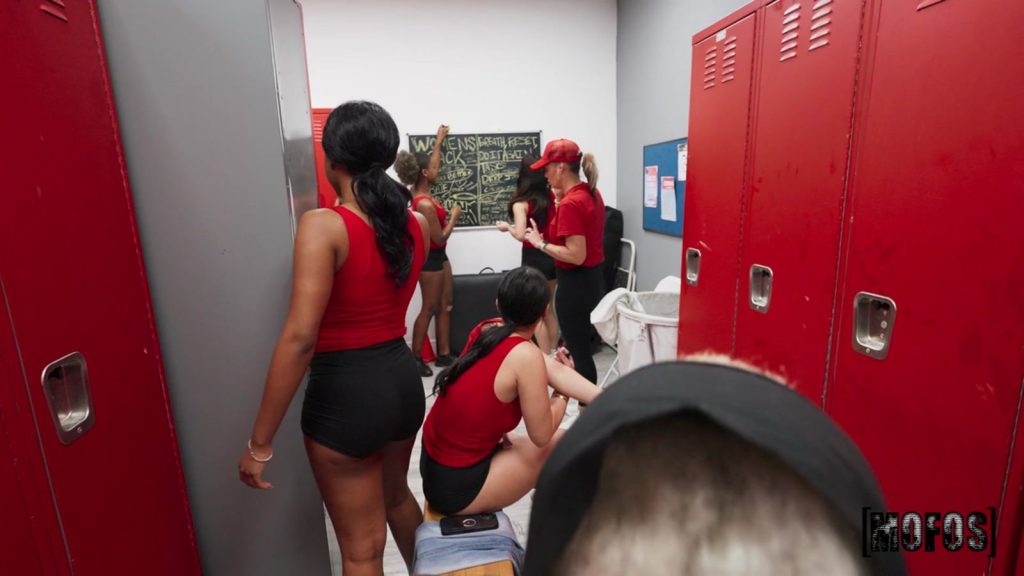 A group of women in red tops and black shorts are in a locker room with red lockers. Some are standing, some are sitting, and a chalkboard with writing is visible on the back wall.