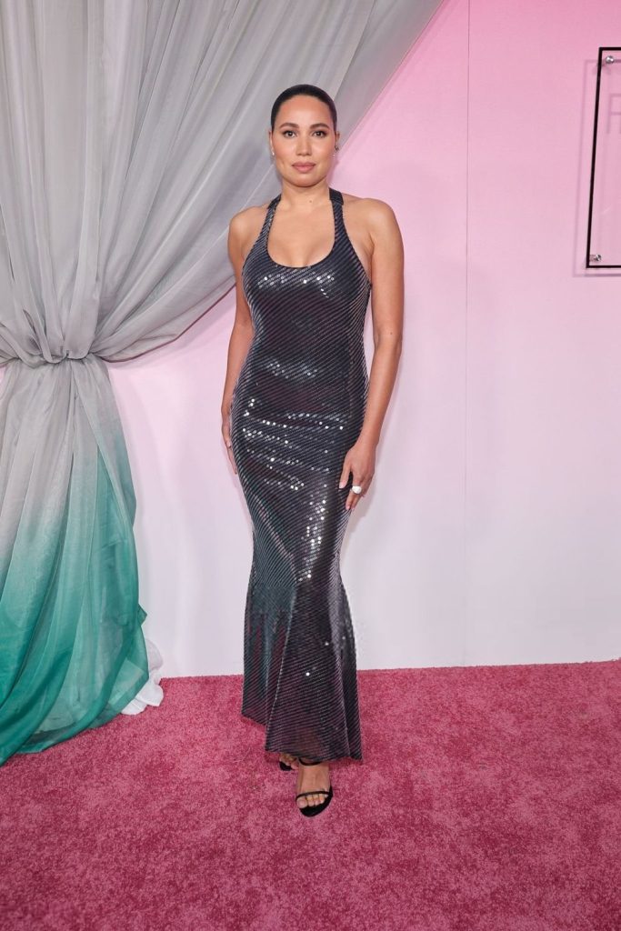 Far away view of a curvy black woman standing on a pink carpet in front of a curtain and pink wall. She is wearing a glittery dress.