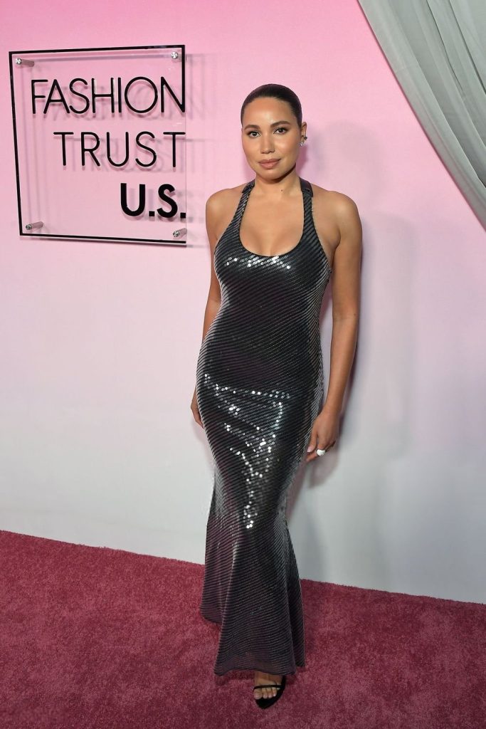 A curvy black woman standing on a pink carpet in front of a pink wall and sign that reads "Fashion Trust USA". She is wearing a glittery dress - Photo #1.