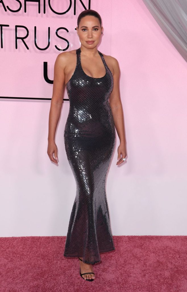A curvy black woman standing on a pink carpet in front of a pink wall and sign that reads "Fashion Trust USA". She is wearing a glittery dress - Photo #2.