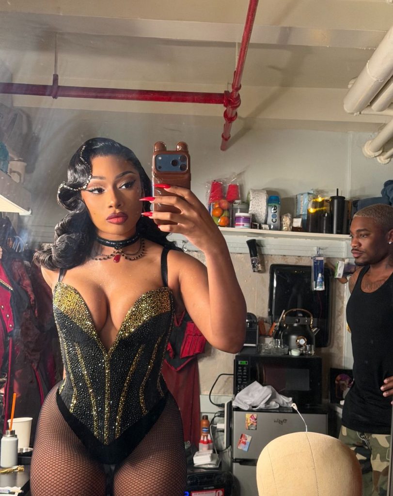 Megan Thee Stallion wearing a corset taking a selfie in a dressing room with various costumes and accessories in the background.