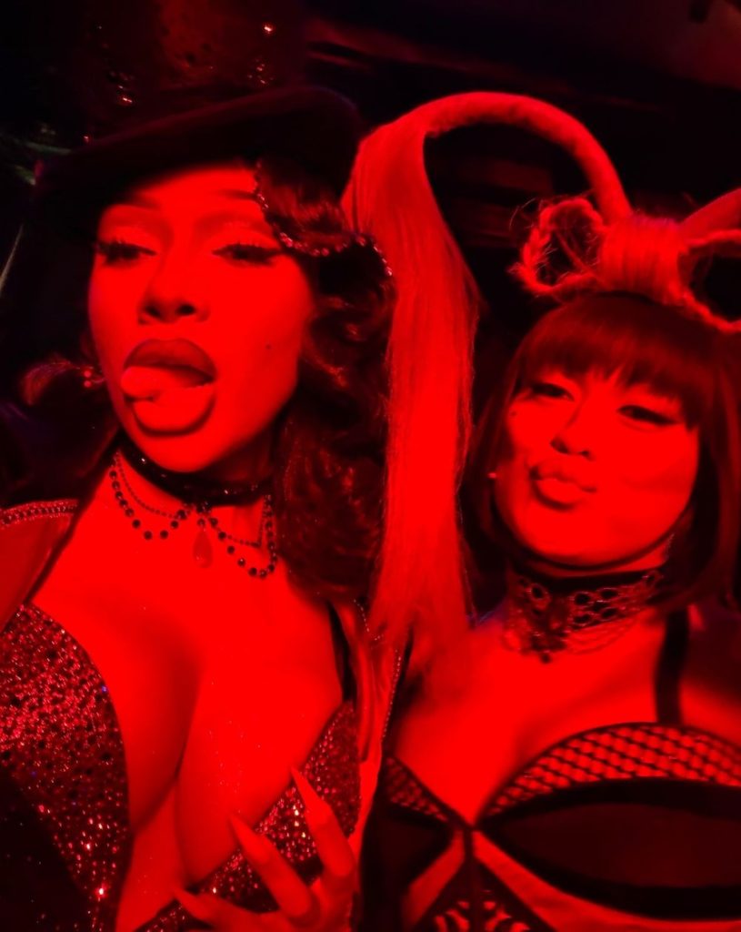Megan Thee Stallion sticks her tongue out when taking a close up selfie with a woman. They are both lit with very red light.