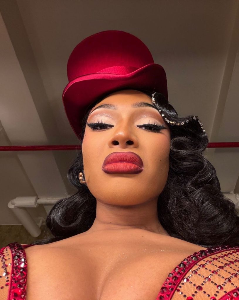 Megan Thee Stallion takes a selfie from below of herself wearing makeup and a red top hat.