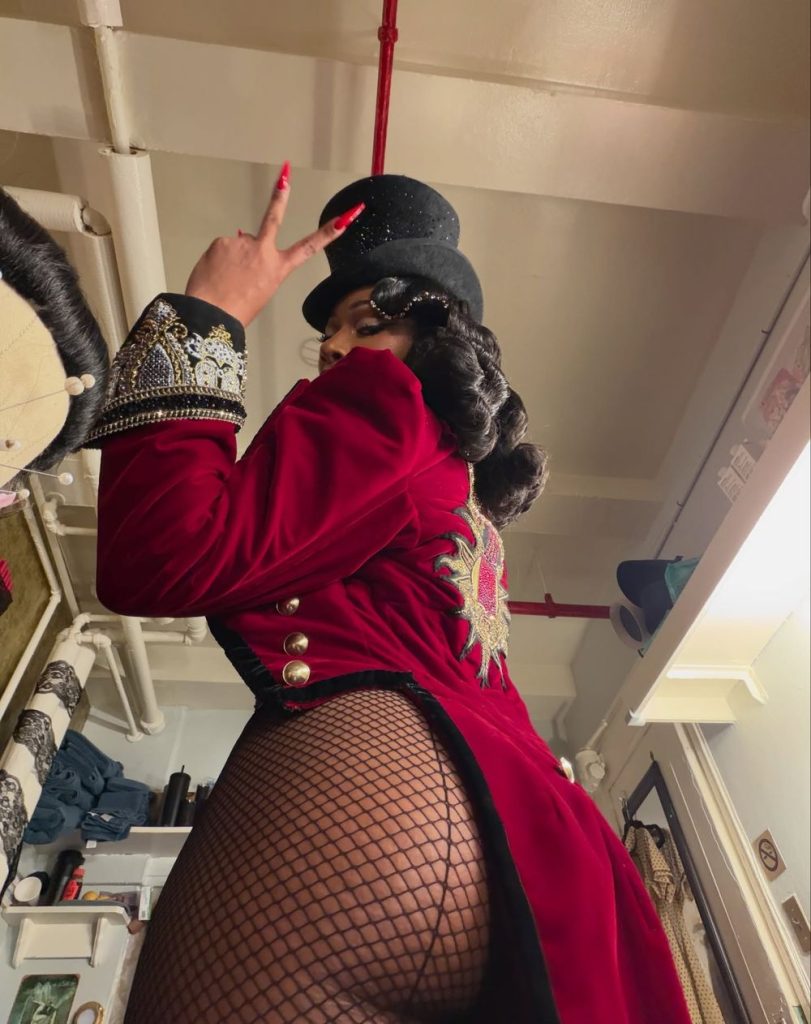 Side view of Megan Thee Stallion wearing fishnet stockings and a top hat. She is dressed like a magician and throws up the peace sign.