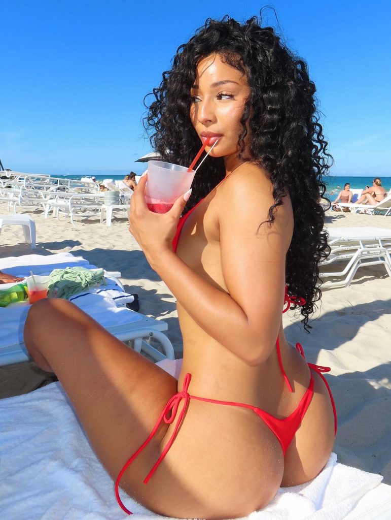 A black woman with long curly hair wearing a red bikini sits on a beach lounge chair, sipping a drink through a straw. The beach is sunny with clear blue sky, and empty lounge chairs are visible in the background.
