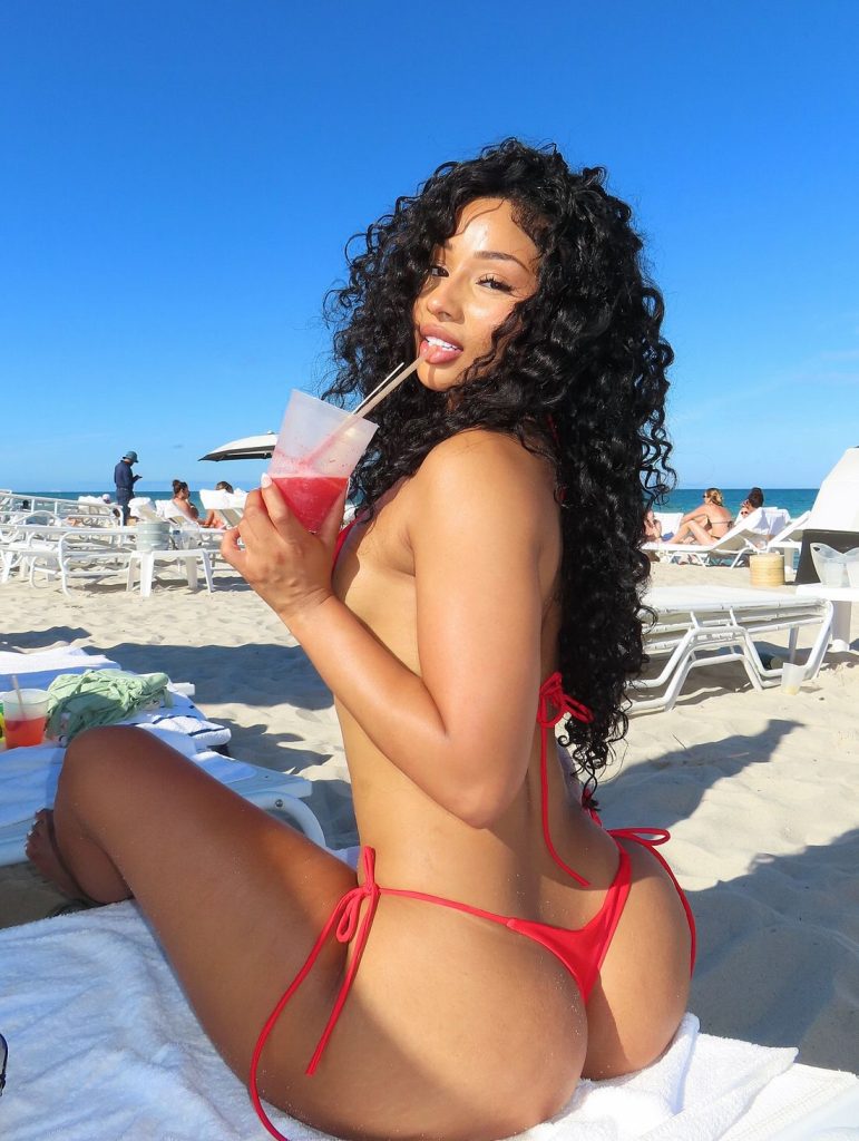 A black woman with long curly hair wearing a red bikini sits on a beach lounge chair, sipping a drink through a straw and looks back towards the camera. The beach is sunny with clear blue sky, and empty lounge chairs are visible in the background.